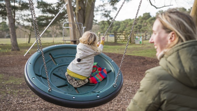 A blonde-haired woman is smiling as her daughter plays on a large swing on the adventure play area at Sutton Hoo